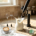 Electric coffee frother mixing milk in a glass jug, with whisk attachments and a coffee cup nearby.